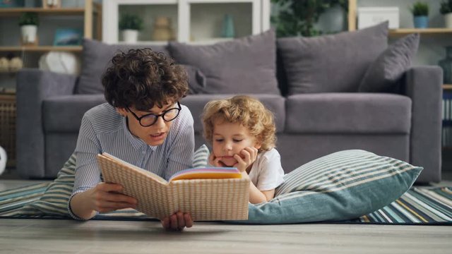 Cute little boy is listening to fairy-tale while his caring mother is reading book lying on floor at home. Parenthood, happy children and interior concept.