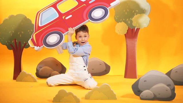 Cheerful little boy plays in the studio with toyful scenery on the yellow background. Happy kid with toyful carton car in his hands.