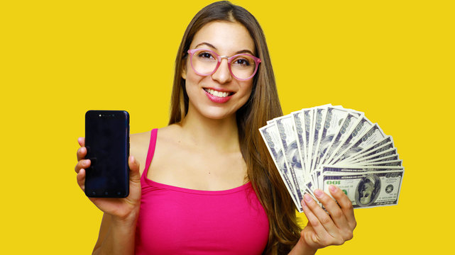 Image of excited young lady isolated over yellow background. Looking camera showing display of mobile phone holding money.