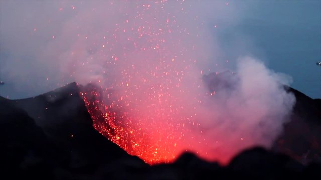 Close up of a lava fountain exploding from a volcano mouth. On the background is the sea with boats
