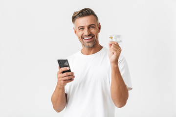 Image of handsome man 30s wearing casual t-shirt holding smartphone and plastic credit card