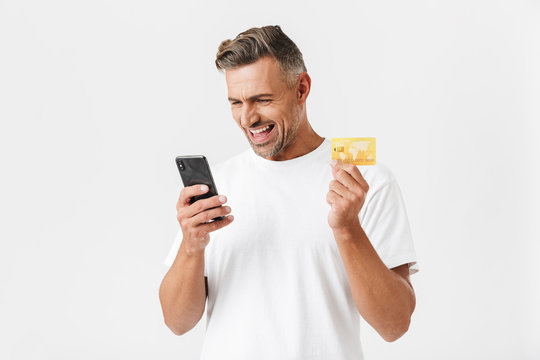 Image of positive man 30s wearing casual t-shirt holding smartphone and plastic credit card