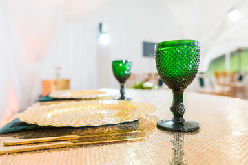 Golden dishes, green wine glasses and napkins. Catering concept. Interior of tent for wedding dinner, ready for guests. Served banquet table.