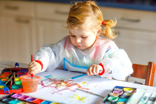 Cute adorable baby girl learning painting with water colors. Little toddler child drawing at home, using colorful brushes. Healthy happy daughter experimenting with colors, water at home or nursery