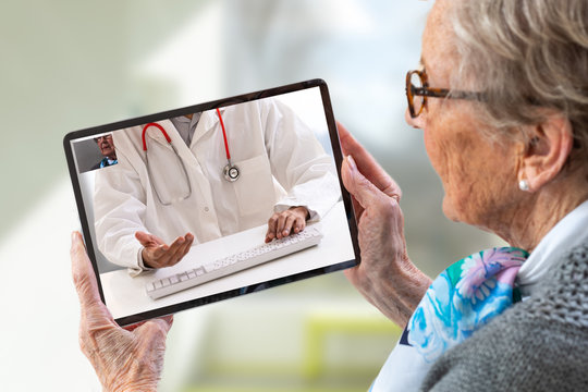 Senior woman consults a e-health doctor with tablet computer sitting in soft chair. In touchscreen, male doctor: With telehealth application patient can reach relevant specialist remotely.