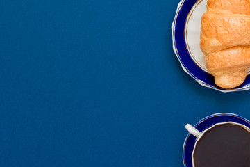 top view of cup of black coffe and croissant on a plate on blue background with copy space. Morning breakfast in french style.