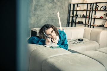 Naklejka premium Tired young dark-haired musician leaning on the couch and writing down