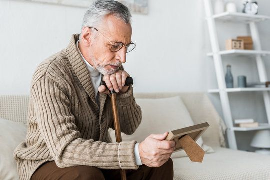 upset retired man looking at photo frame while holding walking stick