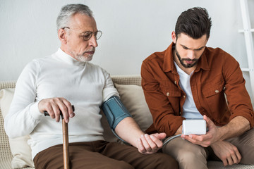 Fototapeta premium bearded man measuring blood pressure of senior father in glasses