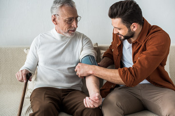 bearded man smiling while measuring blood pressure of happy senior father in glasses