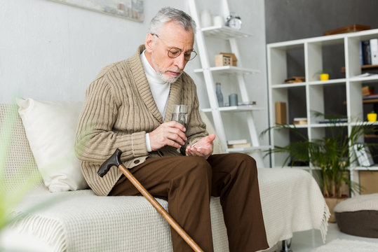 retired man holding pill and glass of water while sitting on sofa near walking cane