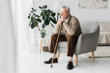 pensive retired man in glasses sitting in armchair at home