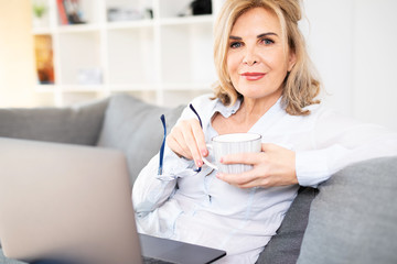 Attraktive reife Frau sitzt zuhause mit Kaffe und Latop auf der Couch 