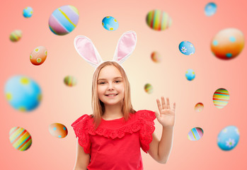 easter, holidays and childhood concept - happy girl wearing bunny ears headband waving hand over colored eggs on living coral background