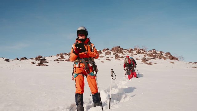 Proud Mountaineer Stands On Snow Hill, Sticking A Ski Pole In The Snow And Looking At A Map Of The Route In The Tablet