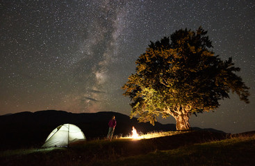Happy female hiker relaxing at summer night camping in the mountains beside campfire, glowing tourist tent and big old tree. Back view of woman enjoying view of night sky full of stars and Milky way.