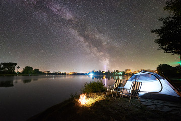 Night camping. Tourist tent, two chairs and campfire on lake shore under amazing evening sky full of stars and Milky way, quiet water surface, city lights on background. Outdoor lifestyle concept