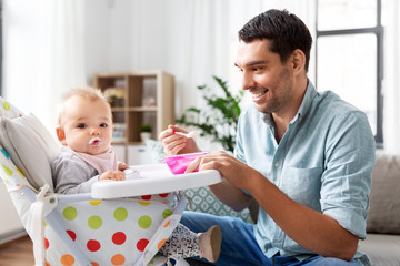 family, food, eating and people concept - happy father feeding little baby daughter sitting in highchair with puree by spoon at home