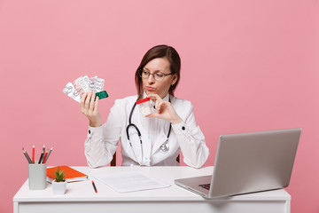 Female doctor sit at desk work on computer with medical document hold pills in hospital isolated on pastel pink wall background. Woman in medical gown glasses stethoscope. Healthcare medicine concept.