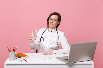 Female doctor sit at desk work on computer with medical document hold water in hospital isolated on pastel pink wall background. Woman in medical gown glasses stethoscope. Healthcare medicine concept.