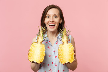 Excited young woman in summer clothes holding halfs of fresh ripe pineapple fruit isolated on pink pastel wall background in studio. People vivid lifestyle relax vacation concept. Mock up copy space.