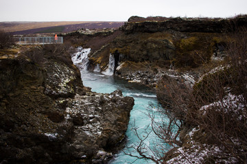 Waterfall view Iceland at wintertime with clear bleu water, person standing in a orange jacket looking at a waterfall. 