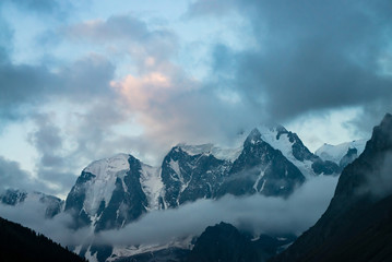 Low cloud before huge glacier. Giant snowy rocky mountains under cloudy sky. Thick mist in mountains at early morning. Impenetrable fog. Cold rocks. Dark atmospheric landscape. Tranquil atmosphere.