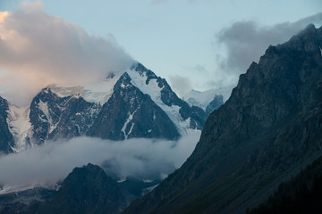 Low cloud before huge glacier. Giant snowy rocky mountains under cloudy sky. Thick mist in mountains at early morning. Impenetrable fog. Cold rocks. Dark atmospheric landscape. Tranquil atmosphere.