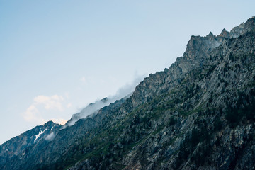 Fototapeta na wymiar Ghostly giant rocks with trees in thick fog. Mysterious huge mountain with snow in mist. Early morning in mountains. Impenetrable fog. Dark atmospheric eerie landscape. Tranquil mystic atmosphere.