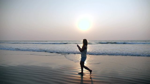 Young beautiful girl cute blonde with loose hair  walking in sunset rays and taking a photo selfie on a tropical sandy beach on sea shore. Silhouette of slim woman with smartphone in ocean waves.