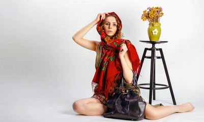 Fashion woman holding black handbag wearing folk dress and red scarf warp on head, sitting on the floor, posing on gray backgroun. Nice girl posing with lady handbag, studio shot