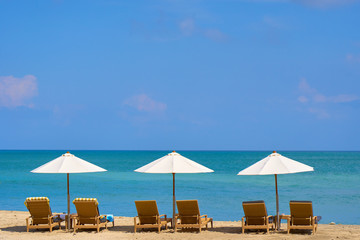 Three white beach umbrellas in a row above lounge chairs on empty sand beach on the ocean shore. Travel and leisure concept. Tropical paradise. Recreation resort.