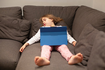 five years old blonde child in pajamas sleeping lying on the coach, with digital tablet on her legs 