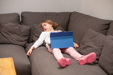 five years old blonde girl sleeping lying on the coach in living room of home, watching digital tablet on her legs 