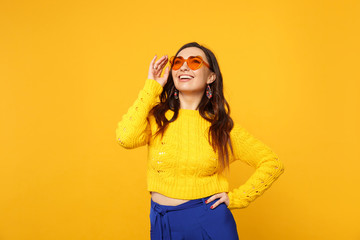 Portrait of joyful young woman in sweater, blue trousers, heart glasses standing, looking up isolated on yellow orange wall background. People sincere emotions, lifestyle concept. Mock up copy space.