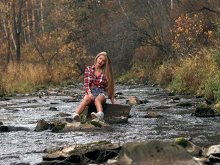 Slim athletic blonde in shorts and a plaid shirt sits in the middle of the river on the stone.
