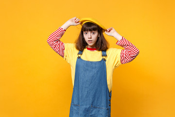 Portrait of perplexed shocked girl teenager in denim sundress keeping hands on french beret isolated on yellow wall background in studio. People sincere emotions lifestyle concept. Mock up copy space.