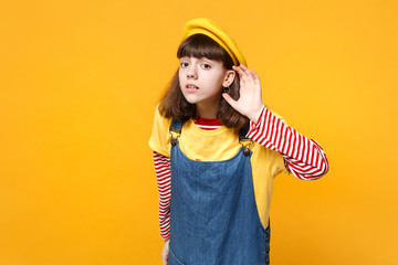 Concerned girl teenager in french beret and denim sundress eavesdrop with hearing gesture isolated...