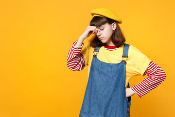 Portrait of tired girl teenager in french beret, denim sundress putting hand on nose, keeping eyes closed isolated on yellow background. People sincere emotions, lifestyle concept. Mock up copy space.