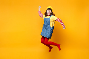 Fototapeta na wymiar Excited girl teenager in french beret, denim sundress spreading hands, jumping and fooling around isolated on yellow wall background in studio. People emotions, lifestyle concept. Mock up copy space.