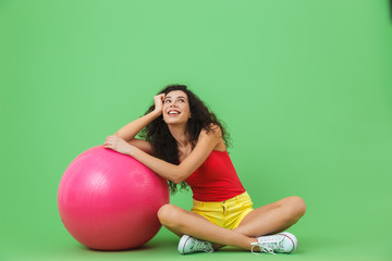 Image of beautiful woman sitting on floor with fitness ball during aerobics against green wall