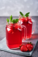Homemade strawberry tea with mint in mason jar on gray concrete table background, copy space. Cold summer berry drink in sparkling glasses. Fresh vitamin beverage