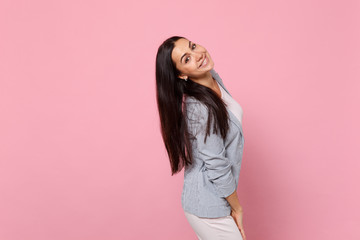 Side view of smiling cheerful young woman in striped jacket standing, looking camera isolated on pink pastel wall background in studio. People sincere emotions lifestyle concept. Mock up copy space.