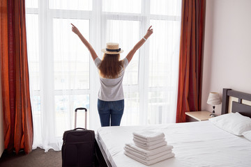 Fototapeta na wymiar Happy young woman staying with raised up arms and luggage in hotel room near big french window, copy space. Back view