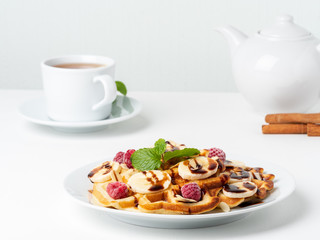 Belgian curd waffles with raspberries, banana, chocolate syrup.  Breakfast with tea on white background, side view