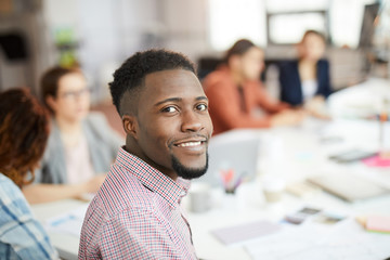 Head and shoulders portrait of young African-American man smiling at camera while sitting at table in business meeting, copy space - obrazy, fototapety, plakaty