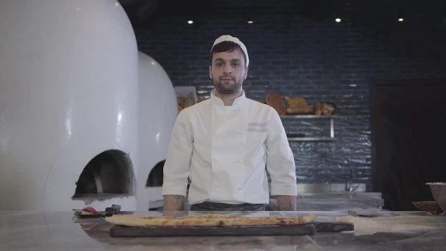 Portrait of chef in white uniform demonstrating freshly baked fatayer lying on the wooden board cutted on pieces in modern restaurant kitchen and looking in camera. Big oven is in the backgound