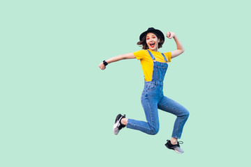 Portrait of happy surprised pretty young hipster girl in blue denim overalls, yellow shirt and black hat jumping and looking at camera and enjoy. indoor studio shot isolated on light green background.