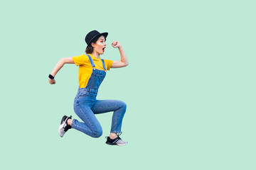 Fototapeta na wymiar Profile side view portrait of surprised funny young hipster girl in blue denim overalls, yellow shirt and black hat jumping in super mario style. indoor studio shot isolated on light green background.