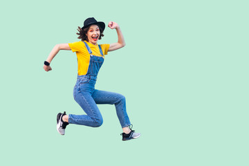 Fototapeta na wymiar Portrait of happy carefree pretty young hipster girl in blue denim overalls, yellow shirt and black hat jumping, looking at camera and enjoying. indoor studio shot isolated on light green background.
